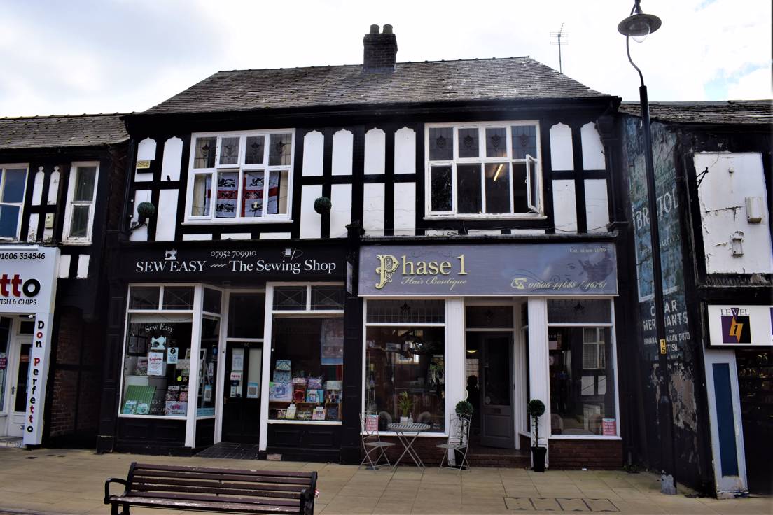 A two-storey building with shop fronts at street level and timber framing with windows above. The shop to the left is Sew Easy, a sewing shop and there is an England flag in the upper window. The shop on the right is a hair boutique called Phase 1. To the right of the image, on the side of the adjacent building, is a ghost advert for Bristol Tipped Cigarettes.